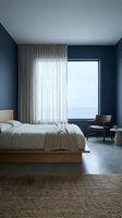 Modern bedroom with navy blue walls, minimalist wooden bed with white linens, floor-to-ceiling sheer curtains, and panoramic window overlooking water. Contemporary interior design for home decor and lifestyle projects.