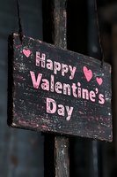 Rustic wooden sign with pink lettering reading Happy Valentine's Day and heart decorations against dark background, showing weathered vintage texture and charm.