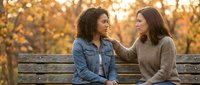 Two diverse young women having intimate conversation on park bench during golden hour sunset with warm bokeh lights creating supportive friendship moment outdoors.
