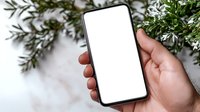 Hand holding modern smartphone with blank white screen against marble background with fresh green branches and white flowers, mockup for app design.
