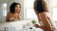 Young African American woman applying skincare routine in modern bathroom mirror reflection for morning beauty regimen and self-care wellness.