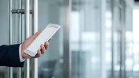 Close-up of male hand holding white smartphone in modern office environment with glass windows and blurred background, suitable for business technology and communication concepts.