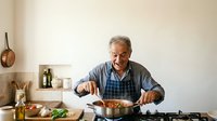 Senior man with gray hair and Caucasian ethnicity, wearing blue checkered shirt and apron, cooking at gas stove in modern kitchen. Suitable for culinary, lifestyle, and home cooking content.