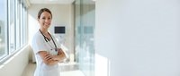 Confident female doctor with stethoscope standing in modern hospital corridor with arms crossed, smiling at camera in bright medical facility.