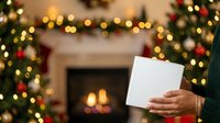 Person holding blank white card with festive holiday decorations, garland, and fireplace with warm lights in background for seasonal greeting or announcement.