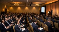 Business conference with diverse professional audience listening to speaker presentation in elegant hotel ballroom with warm lighting and modern decor.