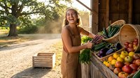 Young blonde woman farmer in apron laughing while arranging fresh organic vegetables and fruits at rustic wooden farm stand during golden hour sunset.
