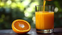 Fresh orange juice being poured into glass with orange slice on wooden table in natural sunlight for healthy breakfast and nutrition concepts.