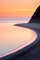 Lone figure stands on curved coastline during peaceful sunset, silhouetted against dramatic cliff and pink-orange sky reflecting in calm ocean waters.