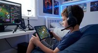 Young male gamer with headset playing mobile game on tablet in modern gaming room setup with LED lights and multiple screens for streaming content.