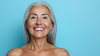 Confident mature Caucasian woman with long silver hair and natural smile against turquoise background showing joy and vitality in aging gracefully.