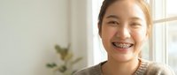 Young Asian woman with dental braces smiling happily in bright modern home interior. Orthodontic treatment and dental care concept for healthcare marketing.