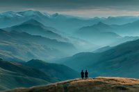 Two hikers standing on mountain ridge overlooking layered blue peaks in misty atmospheric landscape for outdoor adventure and travel concepts.