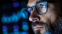 Close up profile of bearded man in glasses looking at financial charts and market data glowing in blue on digital display, focused on stock analysis.