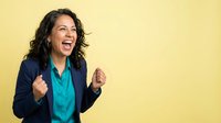 Young woman with dark curly hair and olive complexion wearing navy blazer and teal blouse, smiling enthusiastically with fists raised against yellow background for business and lifestyle content.