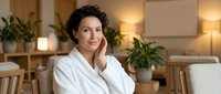 Young man with curly dark hair and olive complexion wearing white bathrobe in modern spa or wellness center with plants and warm lighting, relaxation and self-care concept.