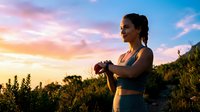 Athletic Asian woman checking fitness tracker during outdoor workout at golden hour sunset with colorful sky and natural landscape background.