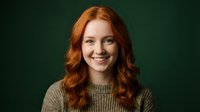 Happy young Caucasian woman with long curly red hair wearing cozy knit sweater smiling at camera against dark green background for lifestyle content.
