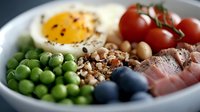 Fresh protein bowl with soft boiled egg, green peas, ham, chickpeas, quinoa, cherry tomatoes and blueberries on white ceramic plate, healthy breakfast.