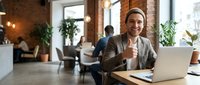 Caucasian male professional in brown blazer and beige shirt smiling at camera while working on laptop in modern loft office with exposed brick, plants, and colleagues collaborating in background.