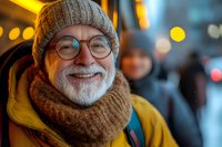 Cheerful senior man with white beard wearing winter knit hat, scarf and glasses smiling outdoors in evening city lights with warm bokeh background.