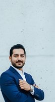 Middle Eastern male professional in navy blazer and white shirt, arms crossed confidently against textured concrete wall. Corporate portrait for business, leadership, and executive use.