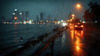 Rainy night city street view through car windshield with water droplets, blurred traffic lights and urban bokeh creating atmospheric moody scene.