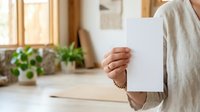 Person in neutral beige linen clothing holding blank white card in bright, minimalist home interior with potted plants and natural window light for mockup.