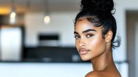 Young South Asian woman with glowing skin and messy bun hairstyle against blurred modern interior background showcases natural makeup and dewy complexion in beauty portrait.
