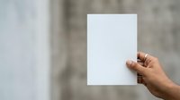 Hand holding blank white paper sheet against concrete wall background for mockup and branding purposes.