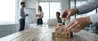Business professional arranging wooden blocks with strategy icons on conference table while team collaborates in modern office background.