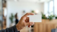 Closeup of male hand holding blank white business card in modern office environment with blurred background and natural lighting for branding and corporate messaging.