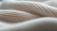 Soft beige knitted fabric texture in macro shot showing detailed wool or cotton yarn structure with gentle waves and folds creating abstract pattern.
