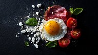 Sunny side up egg with crispy prosciutto, fresh cherry tomatoes and basil leaves on dark background with crumbled feta cheese, dramatic food photography.