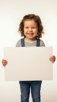 Happy toddler girl with curly brown hair holding blank white sign, smiling joyfully in striped shirt and denim overalls for advertising mockup.
