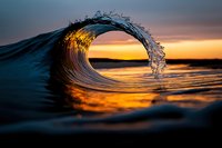 Dramatic ocean wave curling at sunset creates natural frame with golden light reflecting on water surface, capturing powerful motion and serene coastal atmosphere.