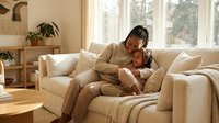 Young African American woman in beige loungewear relaxing on cream sofa with smartphone in bright modern living room with natural light and plants.