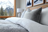 Cozy mountain retreat bedroom with plush white bedding, gray pillows, and stunning snow-capped mountain views through large window.