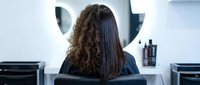 Young woman with curly brown hair receiving professional hair straightening treatment at modern salon with stylist using flat iron for hair transformation.