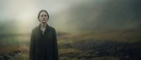 Young woman with fair complexion and light-colored hair standing in misty, desolate moorland landscape wearing dark coat. Moody atmospheric scene for dramatic storytelling and editorial use.