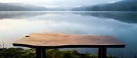 Wooden bench overlooking calm lake with misty mountains and forest reflection, serene nature landscape for travel and outdoor relaxation themes.