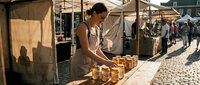 Woman vendor arranging jars of honey or preserves at outdoor market stall. Female merchant selling homemade goods at farmers market or street fair.