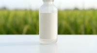 White glass milk bottle with blank label on wooden table against blurred green farm field background for dairy product packaging and agricultural marketing.