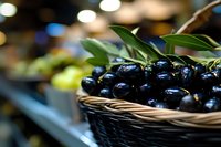 Fresh black olives in wicker basket at grocery store market display with green leaves garnish and blurred produce section background.
