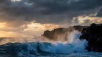 Dramatic ocean waves crashing against dark volcanic rocks under stormy sunset sky with golden light breaking through clouds creating powerful seascape.