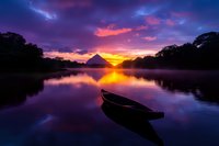 Wooden boat on tranquil lake at dramatic purple sunset, mountain silhouette reflecting in calm water with vibrant orange sky and misty atmosphere.