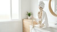 Skeleton wearing white bathrobe and head wrap standing at bathroom sink with potted plant and mirror, bright natural light from window, humorous home decor setup.