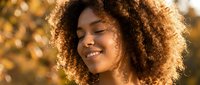 Young African American woman with curly hair smiling peacefully in warm golden sunlight during autumn season outdoors.