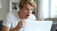 Young blonde man looking shocked and surprised while using laptop computer at home, expressing amazement at screen content with open mouth and wide eyes.