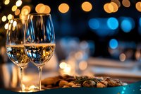 Two elegant wine glasses filled with white wine on table with appetizers and warm bokeh lights creating romantic dining atmosphere.